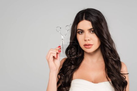 Téléchargez les photos : Pretty brunette woman with long and healthy hair holding scissors and looking at camera isolated on grey - en image libre de droit