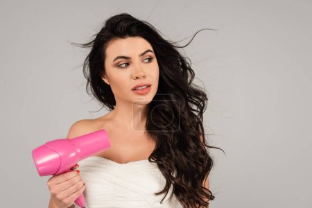 brunette woman in white top holding hair dryer and looking away isolated on grey