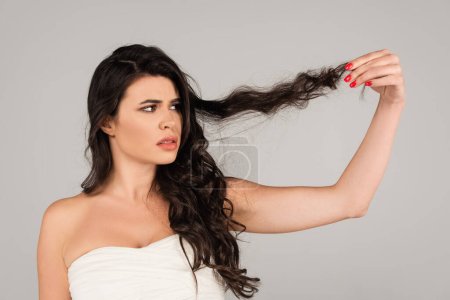 displeased woman in white top looking at damaged and tangled hair isolated on grey