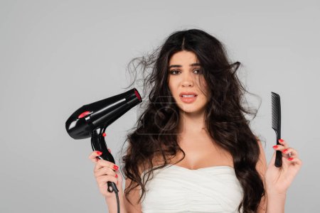 upset woman with damaged and tousled hair holding comb and hair dryer while looking at camera isolated on grey