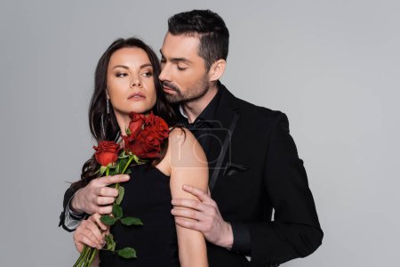 bearded man in black formal wear holding red roses near brunette woman isolated on grey 