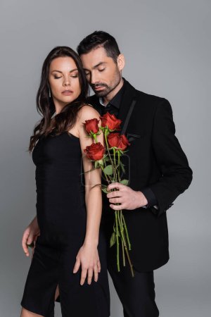 bearded man in suit holding red roses near brunette woman isolated on grey