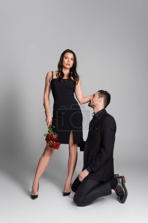 full length of dominant woman in black dress holding red roses near bearded man in suit on grey 