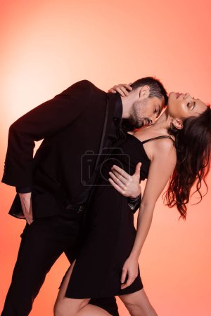 Foto de Passionate and bearded man kissing chest of seductive woman in black dress on pink - Imagen libre de derechos