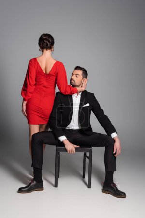 Foto de Elegant woman in dress touching face of boyfriend sitting on chair on grey background - Imagen libre de derechos