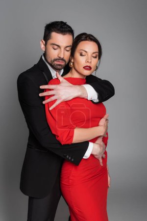 Bearded man in suit hugging seductive woman with burgundy lips isolated on grey 