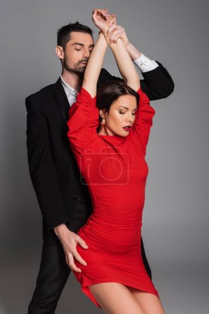 Bearded man touching hands and hip of sexy girlfriend in red dress isolated on grey 