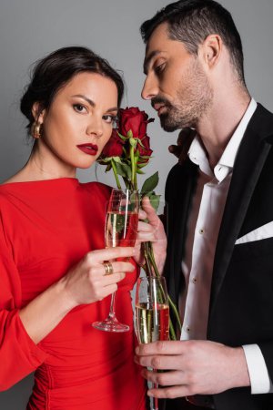 Elegant woman looking at camera near boyfriend with champagne and roses isolated on grey 