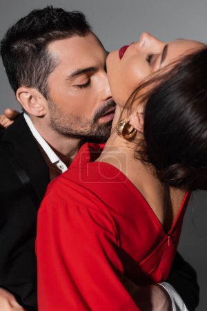 Elegant man in suit kissing neck of sensual girlfriend on grey background 