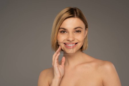 happy woman with naked shoulders looking at camera while smiling isolated on grey 