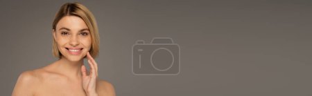 happy woman with naked shoulders looking at camera while smiling isolated on grey, banner 