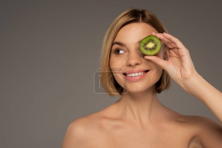 happy young woman with bare shoulders covering eye with kiwi fruit isolated on grey 