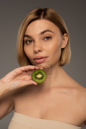 blonde young woman with bare shoulders holding kiwi fruit and looking at camera isolated on grey 