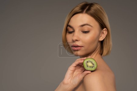 young blonde woman with bare shoulders holding kiwi fruit isolated on grey 