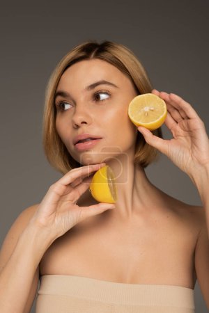 young blonde woman with bare shoulders holding lemon halves and looking away isolated on grey 