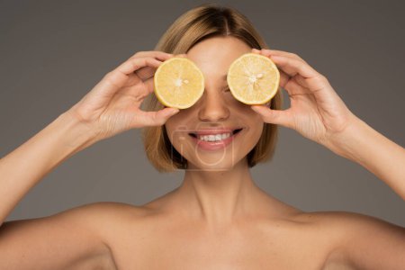 Photo for Happy young woman with bare shoulders covering eyes with lemon halves isolated on grey - Royalty Free Image