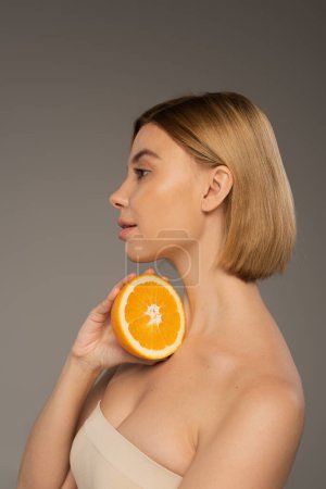 side view of pretty young woman with bare shoulders holding orange isolated on grey 