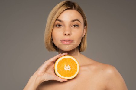 young blonde woman with bare shoulders holding tasty orange and looking at camera isolated on grey 