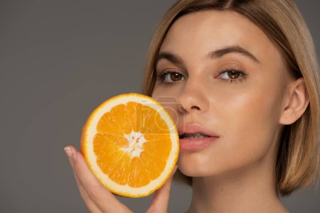 portrait of young blonde woman holding juicy orange half isolated on grey 