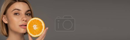 portrait of young blonde woman holding juicy orange half isolated on grey, banner 