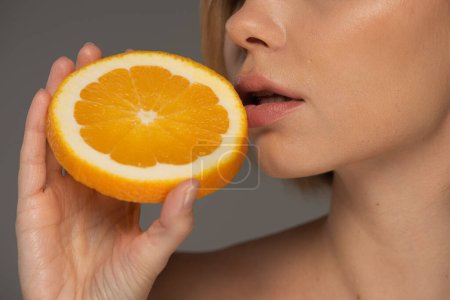 cropped view of young woman holding juicy orange half near soft lips isolated on grey 