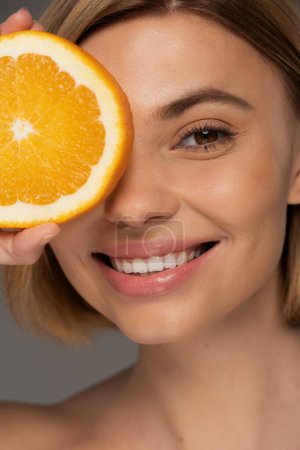 portrait of cheerful blonde woman holding juicy orange half near face isolated on grey 