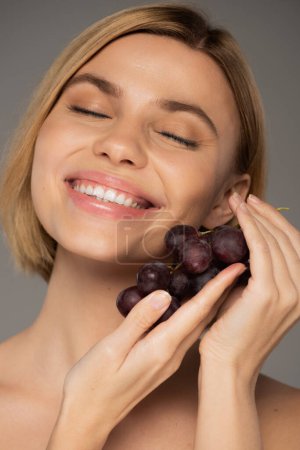 cheerful young woman holding fresh grapes and smiling isolated on grey 