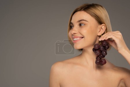 happy young woman with bare shoulders holding ripe grapes near ear isolated on grey 