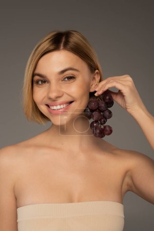 joyful young woman with bare shoulders holding ripe grapes near ear isolated on grey 