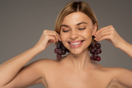 smiling young woman with bare shoulders holding sweet grapes near ears isolated on grey 