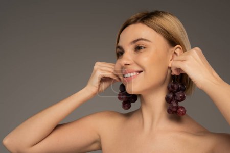 joyful young woman with bare shoulders holding tasty grapes near ears isolated on grey 