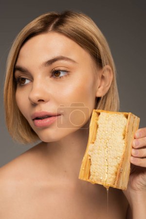 blonde young woman holding honeycomb and looking away isolated on grey 