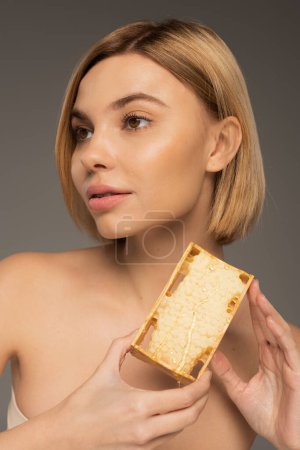 Téléchargez les photos : Blonde young woman with bare shoulders holding honeycomb while looking away isolated on grey - en image libre de droit