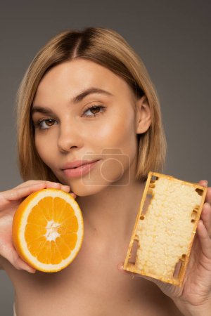 Photo for Blonde young woman holding honeycomb and orange while looking at camera isolated on grey - Royalty Free Image