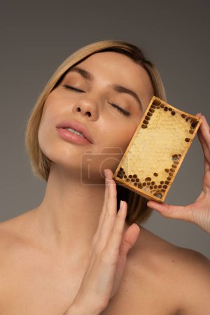 blonde young woman with closed eyes holding wooden frame with sweet honey isolated on grey 