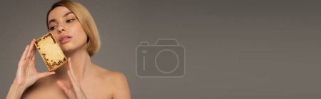 Photo for Blonde young woman holding wooden frame with sweet honey while looking away isolated on grey, banner - Royalty Free Image