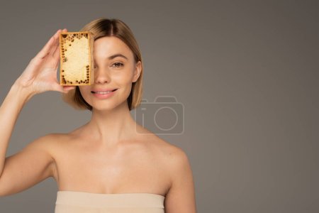 cheerful woman covering eye with honeycomb in wooden frame isolated on grey 
