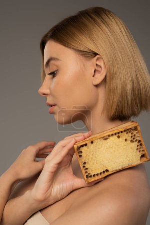 Photo for Side view of blonde woman holding honeycomb in wooden frame isolated on grey - Royalty Free Image