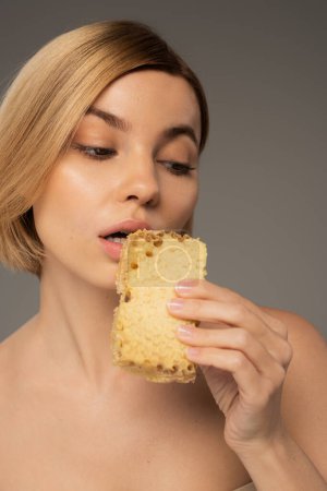 young woman holding piece of sweet honeycomb near mouth isolated on grey