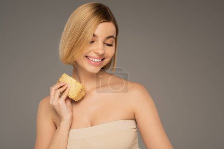cheerful young woman with bare shoulders holding piece of sweet honeycomb isolated on grey