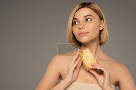 blonde woman with bare shoulders holding piece of sweet honeycomb while looking away isolated on grey