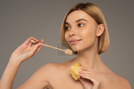 Photo for Pleased woman with bare shoulders holding sweet honeycomb and wooden dipper isolated on grey - Royalty Free Image
