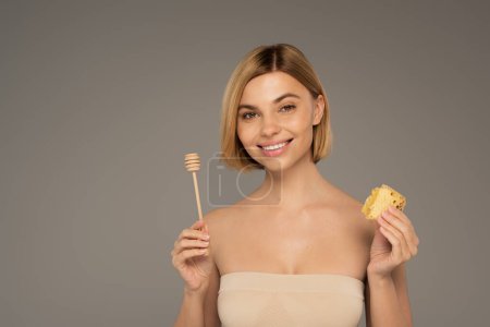 Photo for Happy woman with naked shoulders holding sweet honeycomb and wooden dipper isolated on grey - Royalty Free Image