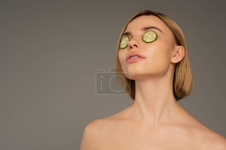 blonde young woman with naked shoulders and fresh cucumbers on eyes isolated on grey 