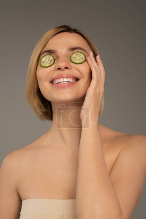 joyful young woman with naked shoulders and fresh cucumbers on eyes isolated on grey 