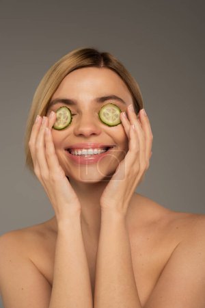 cheerful young woman with naked shoulders and fresh cucumbers on eyes isolated on grey 