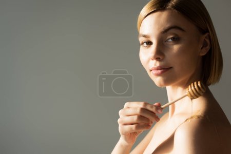 blonde woman holding dipper with sweet honey near bare shoulder isolated on grey