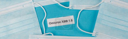 Top view of omicron xbb lettering near protective masks on blue background, banner 