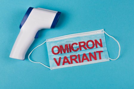 Top view of non-contact pyrometer near medical mask with omicron variant lettering on blue background 