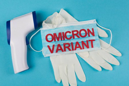 Top view of medical mask with omicron variant lettering near latex gloves and pyrometer on blue background 
